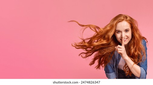 A young woman with long, vibrant red hair flows to the side as she places one finger to her lips, signaling for silence on pink background, copy space - Powered by Shutterstock