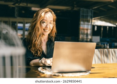 A young woman with long wavy hair and glasses is working on a laptop while sitting at a wooden table in a cozy café. She is smiling and appears relaxed and focused. 