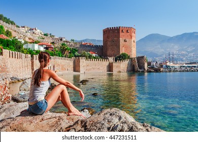 Young woman look at Kizil Kule tower in Alanya peninsula, Antalya district, Turkey, Asia. Famous tourist destination with high mountains. Part of ancient old Castle. Summer bright day