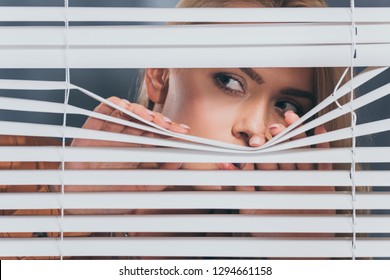 young woman looking away and peeking through blinds, mistrust concept 