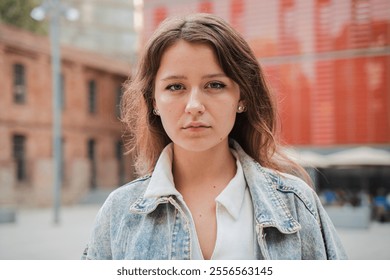 Young woman looking at camera outdoors, wearing casual denim jacket. Teenage girl showcasing a confident and serious expression looking at camera in an urban environment with natural light. High - Powered by Shutterstock