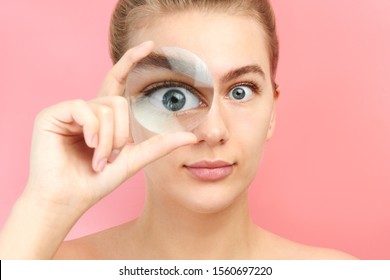 Young Woman Looking At The Camera Through Magnifier With Funny Surprised Expression, Closeup Isolated On Pink. Enlarged Eye, Optical Illusion, Search