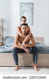 Young Woman Looking At Camera While Embracing Shirtless Man Sitting On Bed In Pajama Pants