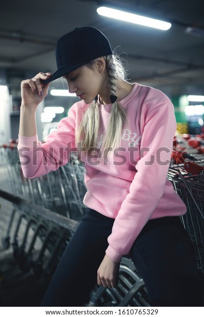 Young woman looking down and touch black cap.
Shopping carts - supermarket underground car park. Street style
fashion.