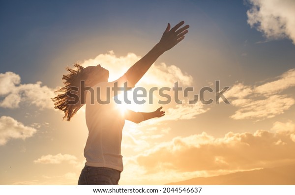 Young woman looking up feeling energized by the\
warm rays of sunshine lifting arms up to the sunset sky. Letting go\
of your fears concept. 