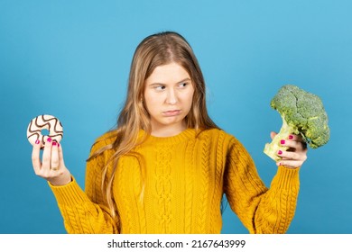 Young Woman Looking With Frustration At Brocoli And Holding Chocolate Donut Isolated On Blue Background.