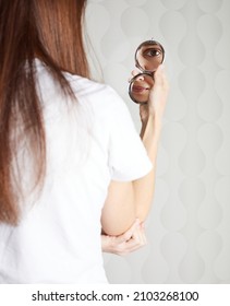 Young Woman Looking In Hand Mirror. Her Back Is Turned To The Camera And Part Of Her Face Reflects In Mirror. Portrait Of Girl With Fresh Healthy Skin