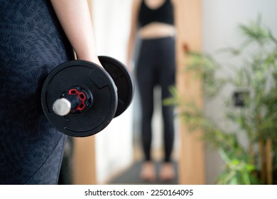 Young woman looking at herself in the mirror while lifting dumbbell in the bedroom. Focus on the dumbbell - Shutterstock ID 2250164095
