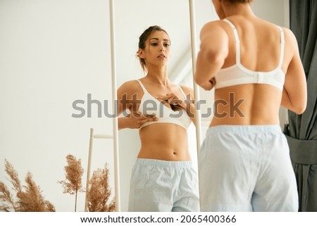 Young woman looking herself in a mirror while doing breast self-exam at home. 