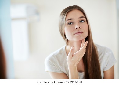 Young Woman Looking At Mirror At Home And Applying Cream On Her Face