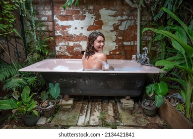 A Young Woman Looking Over Her Shoulder Smiles Happily While Sitting In An Outdoor Bubble Bath In A Lush Tropical Garden.  