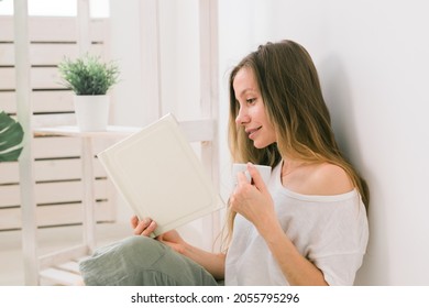 Young Woman Looking Photo Album At Home And Drink Tea. Memories And Leisure Concept.