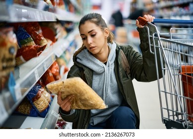 Young woman looking at the price on pasta package while shopping in grocery store.