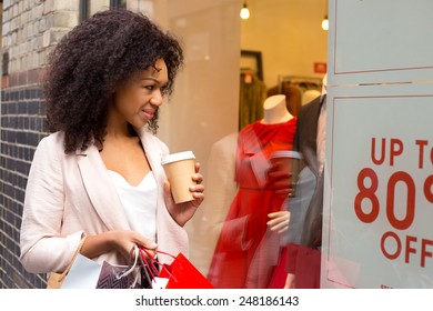 Young Woman Looking In A Shop Widow With A Coffee And Shopping Bags.