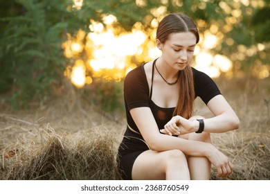 Young woman looking at the smart clock checking pulse sitting on the roadside in forest park. Girl training at sunset on nature, health and sport concept - Shutterstock ID 2368760549