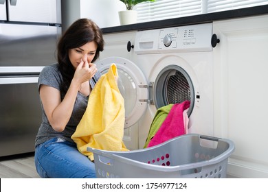 Young Woman Looking At Smelly Clothes Out Of Washing Machine In Kitchen