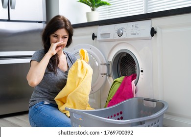 Young Woman Looking At Smelly Clothes Out Of Washing Machine In Kitchen