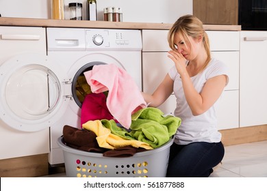 Young Woman Looking At Smelly Clothes Out Of Washing Machine In Kitchen