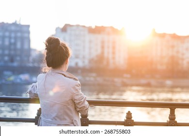 A young woman is looking at the sunset over a river in the city Prague with the old buildings in the background. Soft spring backlit. Color toned image.