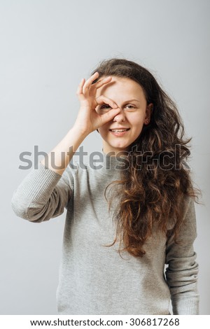 Young woman looking through her fingers, a gesture okay around the eye. On a gray background.