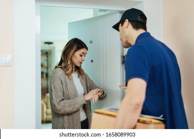 Young Woman Looking At Time On Wristwatch And Feeling Dissatisfied About Late Home Delivery From A Courier. 