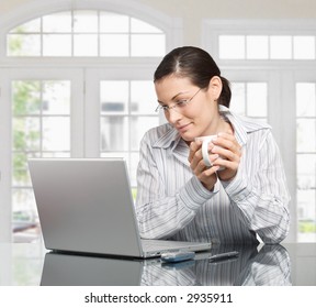 Young Woman Looks At Her Laptop Computer And Drinks Her Coffee. It Is Early Morning In A Light And Clean Home Interior Dominated By White And Soft Tones.