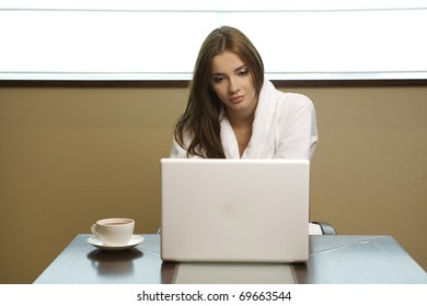 Young Woman Looks At Her Laptop Computer And Drinks Her Coffee. It Is Early Morning In A Light And Clean Home Interior Dominated By White And Soft Tones.