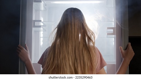 A young woman looks into an open refrigerator, where light shines and fog comes from. Extreme cooling and coolness - Shutterstock ID 2235290463