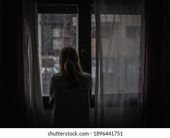 Young woman looks out a window between the curtains while it is snowing.