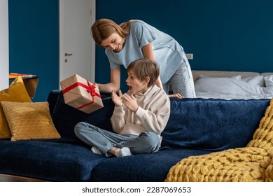 Young woman loving mother surprising son with birthday gift at home, mom giving wrapped box to excited little boy, congratulating child. Overjoyed happy kid sitting on sofa getting present from mum - Shutterstock ID 2287069653
