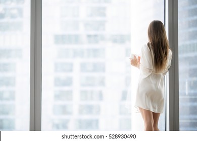Young woman in luxury modern apartment, wearing silk dressing gown drinking coffee and looking at rainy cityscape, enjoying new floor to ceiling windows, dreaming of success in the big city. Rear view