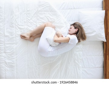 Young Woman Lying In Bed And Holding On To Sore Stomach Top View. Painful Menstruation Concept