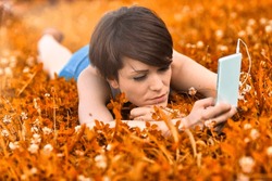 Young Woman Lying In A Meadow Selecting A Tune On Her Mobile Phone As She Spends A Relaxing Day Listening To Music