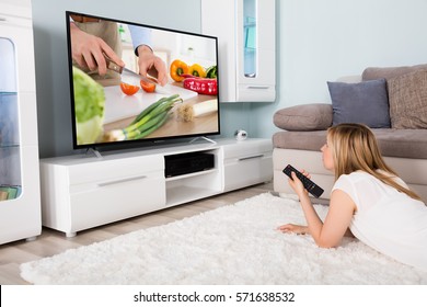 Young Woman Lying On Carpet Watching Cooking Show On Television At Home