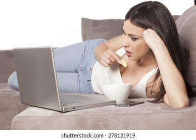 Young Woman Lying On The Couch With A Laptop In Front Of Her And Eating Potato Chips