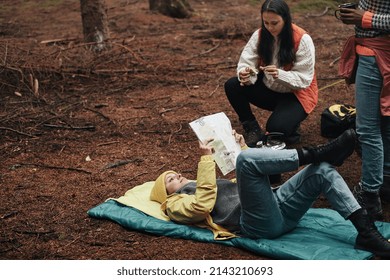 Young Woman Lying On Her Sleeping Bag And Reading A Map While Camping With Friends In The Woods