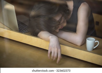 Young woman lying on her arms on the table in cafe in front of laptop with cup of coffee, sleepy, tired, overworked or lazy to work early in the morning