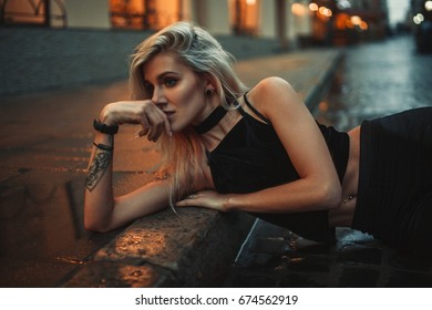 Young Woman Lying On Wet Pavement On City Street In Evening. In Background There Are City Lights. 