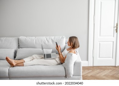 Young woman lying relaxed with phone and laptop on the comfortable couch at home. Front view. Working or leisure time at home