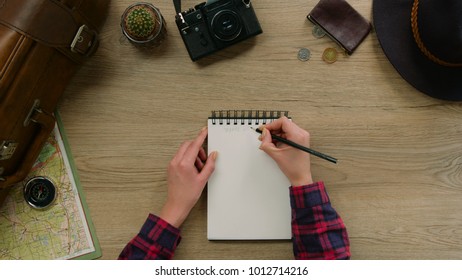 Young Woman Make Some Notes In Note Book On Wood Background. Woman Hands Make Travel List For The Future Trip. Top View.