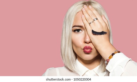 Young woman makes air kiss with her lips and closes one eye by hand. Blonde hair model in white shirt closed her eye with hand. Concept of fun and merriment. Studio portrait on pink background.