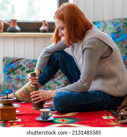 Young Woman Makes Coffee At Home. Sitting On An Oriental Rug, Using A Manual Coffee Grinder To Make Coffee And A Beautiful Cup. Close Up.