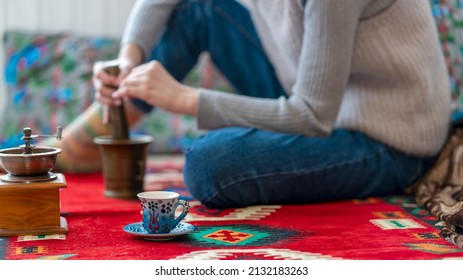 Young Woman Makes Coffee At Home. Sitting On An Oriental Rug, Using A Manual Coffee Grinder To Make Coffee And A Beautiful Cup. Close Up.