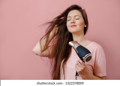 young woman makes hair volume with  hairdryer in hand on pink background