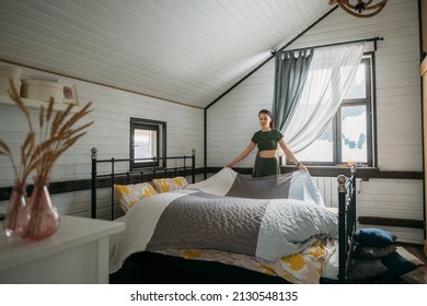 A Young Woman Makes Her Bed In A Cozy Bedroom At Home. A Girl In Home Clothes Does House Cleaning, Changes Bed Linen, Straightens A Blanket On A Large Bed In A Bright Room.