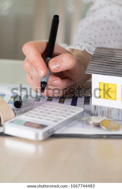 Young woman makes some calculations.\
Small paper house, coins, car in the background.\
Closeup