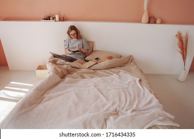 Young woman making notes in bed while working on her laptop. Working from home. Cozy bedroom. Self-isolation.