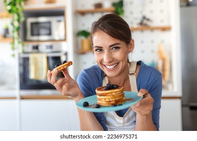 Young woman making pancakes at kitchen. Young housewife enjoying blueberry pancakes for breakfast - Powered by Shutterstock