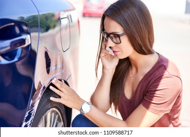Young woman making phone call to insurance agent while inspecting scratched car