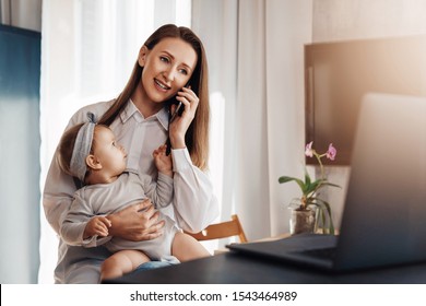 Young Woman Making Phone Call On Her Mobile, Baby Daughter Sitting On Knees And Watching At Mom. Mother Working On Laptop Remotely While Taking Care Of Child At Home. Freelance Career For Young Moms 